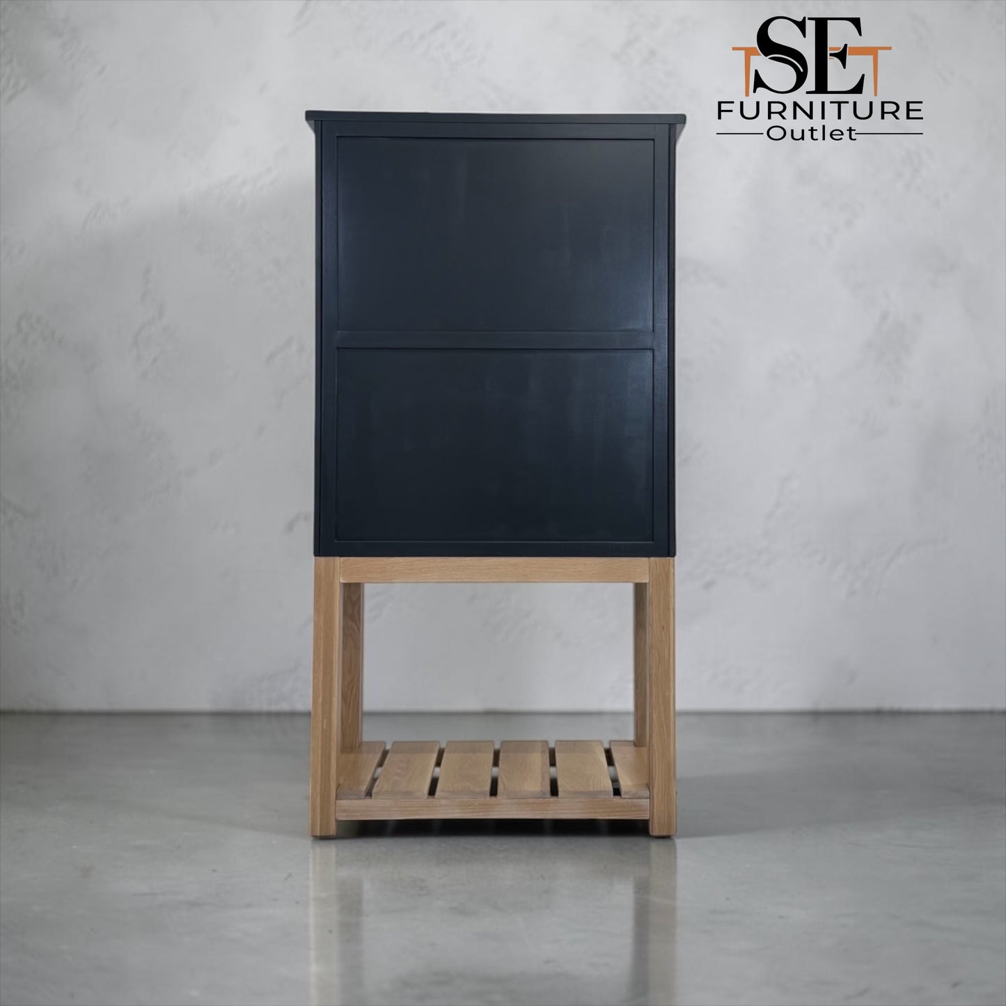A tall black cabinet with a light natural wood base and open slatted shelf, photographed against a light gray textured wall and floor. The SE Furniture Outlet logo is visible in the top right corner.