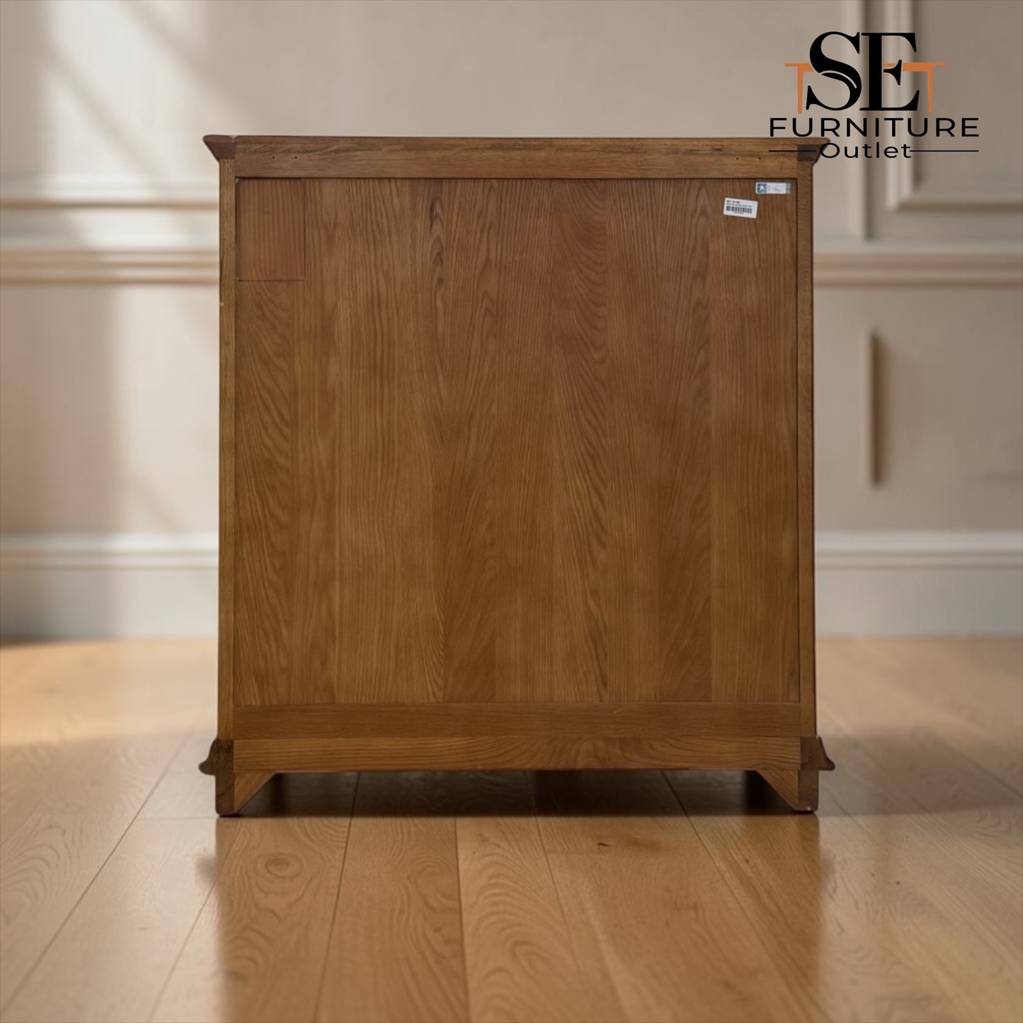 A wooden side cabinet with a warm medium-brown finish and visible wood grain, shown standing on a light wood floor against an off-white paneled wall. A small white price tag is visible on the upper right side, and the 'SE Furniture Outlet' logo appears in the top right corner.