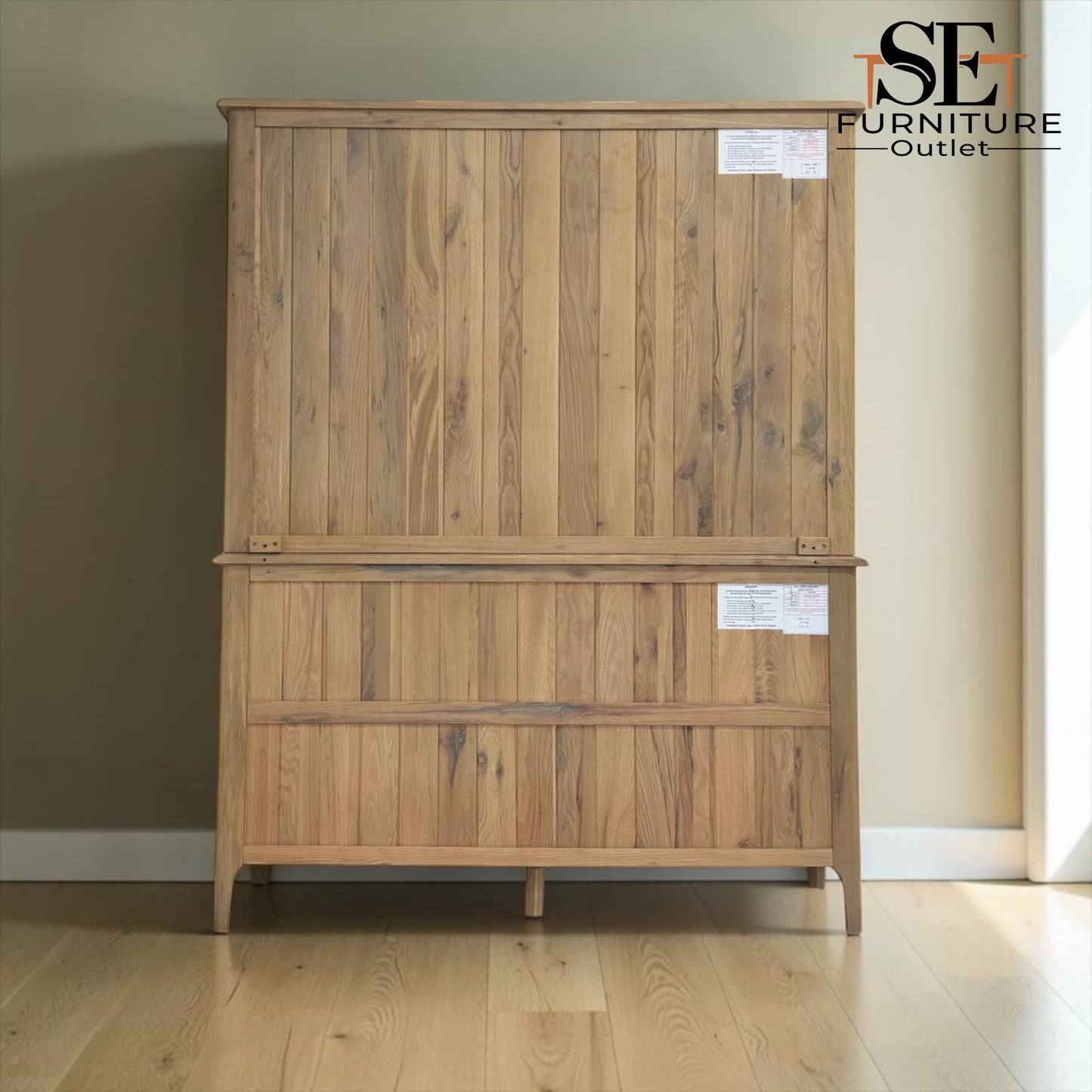 A tall wooden storage cabinet with two doors and two drawers, made from natural oak wood with visible grain and knots, standing on a light wood floor against a beige wall. An 'SE FURNITURE Outlet' logo is visible in the top right corner.