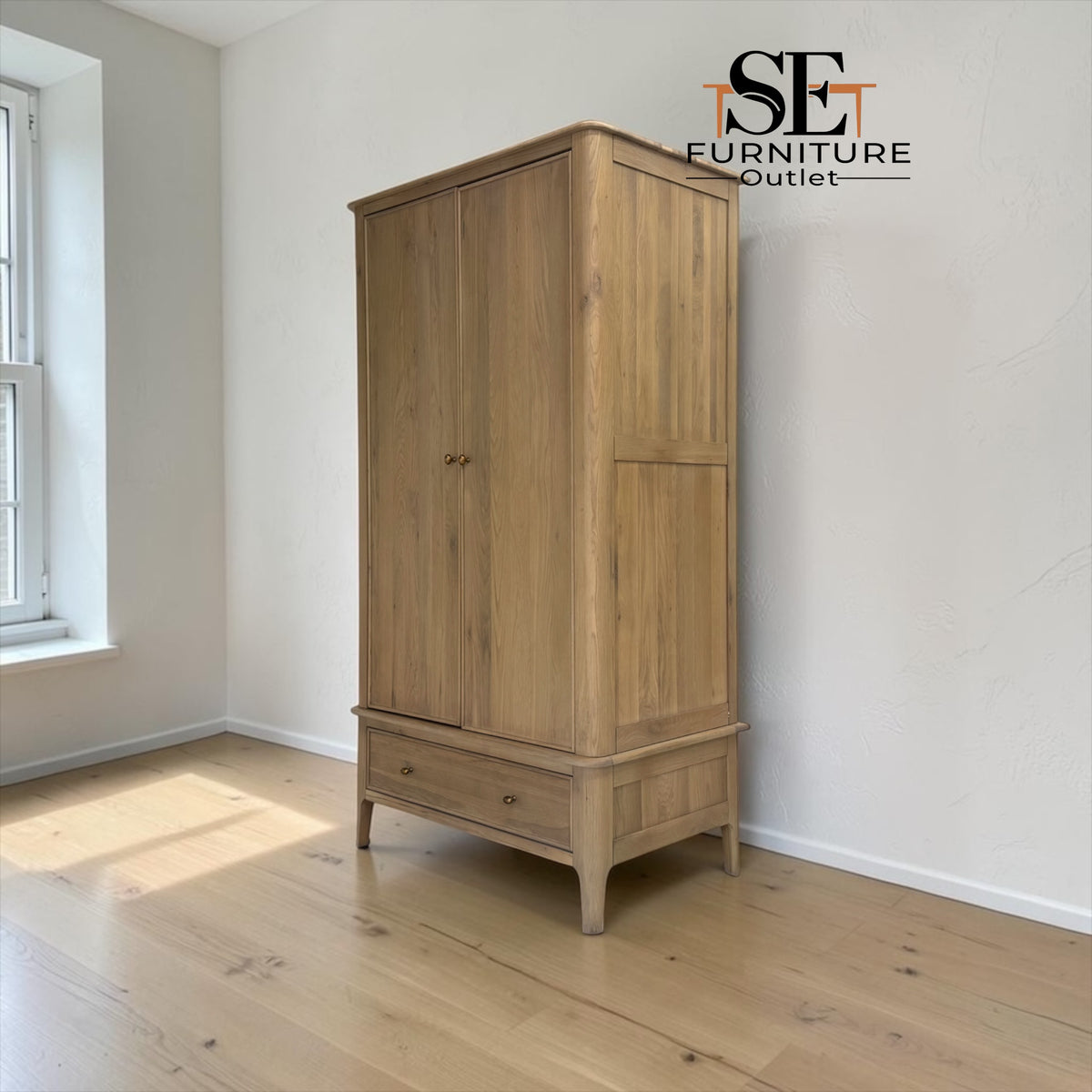 A tall light oak wood armoire cabinet with two doors and a single drawer on a pedestal base, shown in a minimalist room with white walls and light wood floors. The SE Furniture Outlet logo is visible in the top right corner.

