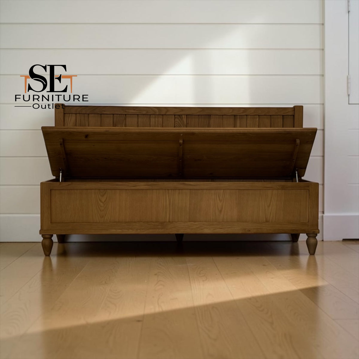 A wooden storage bench with its lid open, set against a white shiplap wall on a light wood floor. The brand logo 'SE FURNITURE OUTLET' is visible in the top left corner.

