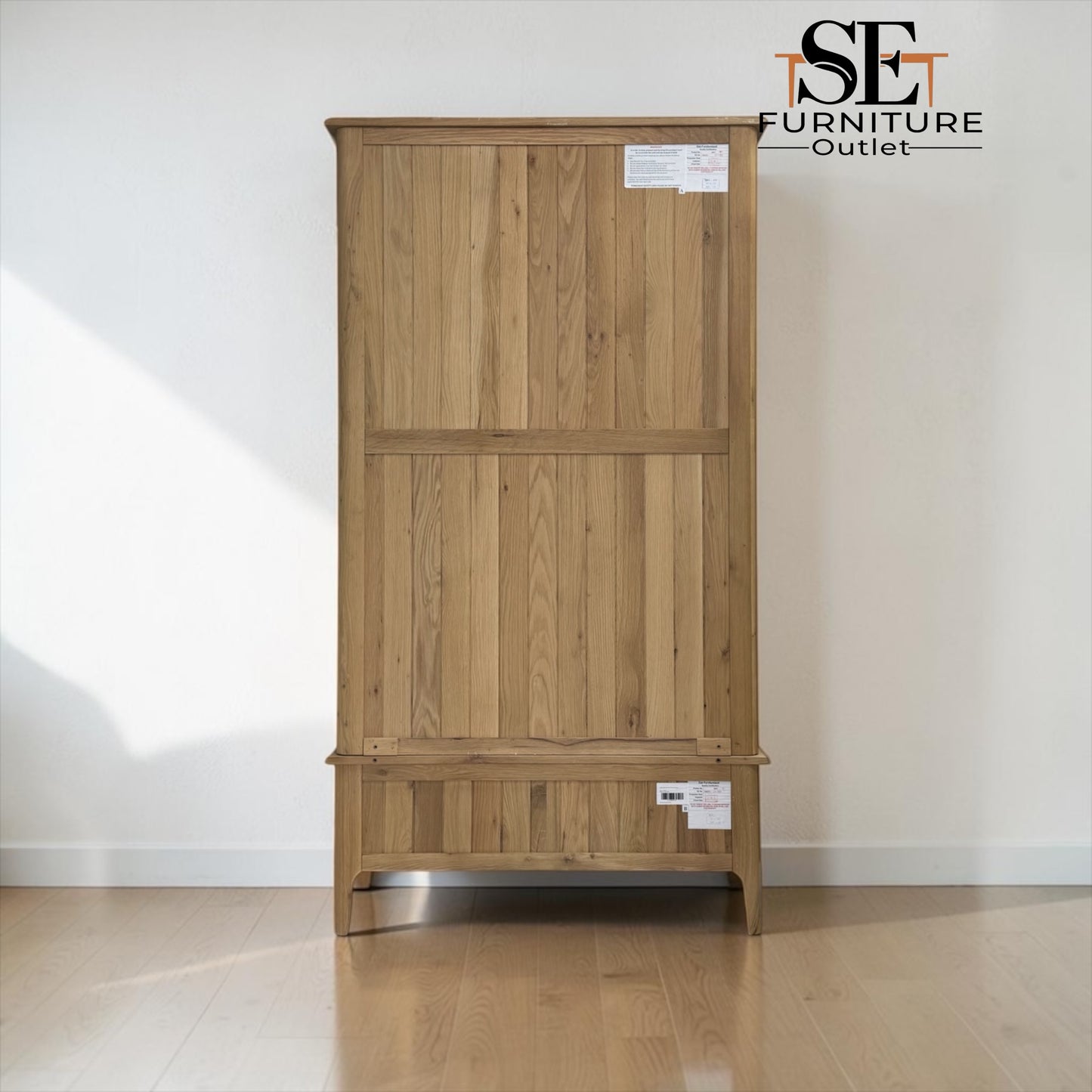 A tall wooden wardrobe or cabinet with a natural oak finish, featuring two upper doors and a lower drawer, shown against a plain white wall on a wooden floor. An 'ISE Furniture Outlet' logo is visible in the top right corner.


