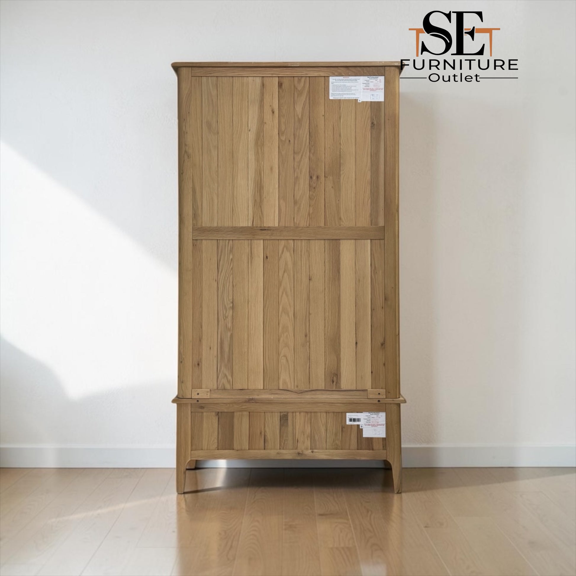 A tall wooden wardrobe or cabinet with a natural oak finish, featuring two upper doors and a lower drawer, shown against a plain white wall on a wooden floor. An 'ISE Furniture Outlet' logo is visible in the top right corner.

