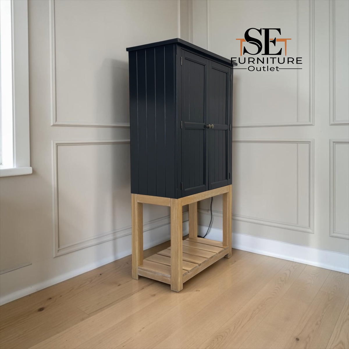 A tall black cabinet with double doors on a light natural wood base, photographed in a room with paneled walls and wood flooring. An 'SE Furniture Outlet' logo is visible in the upper right.

