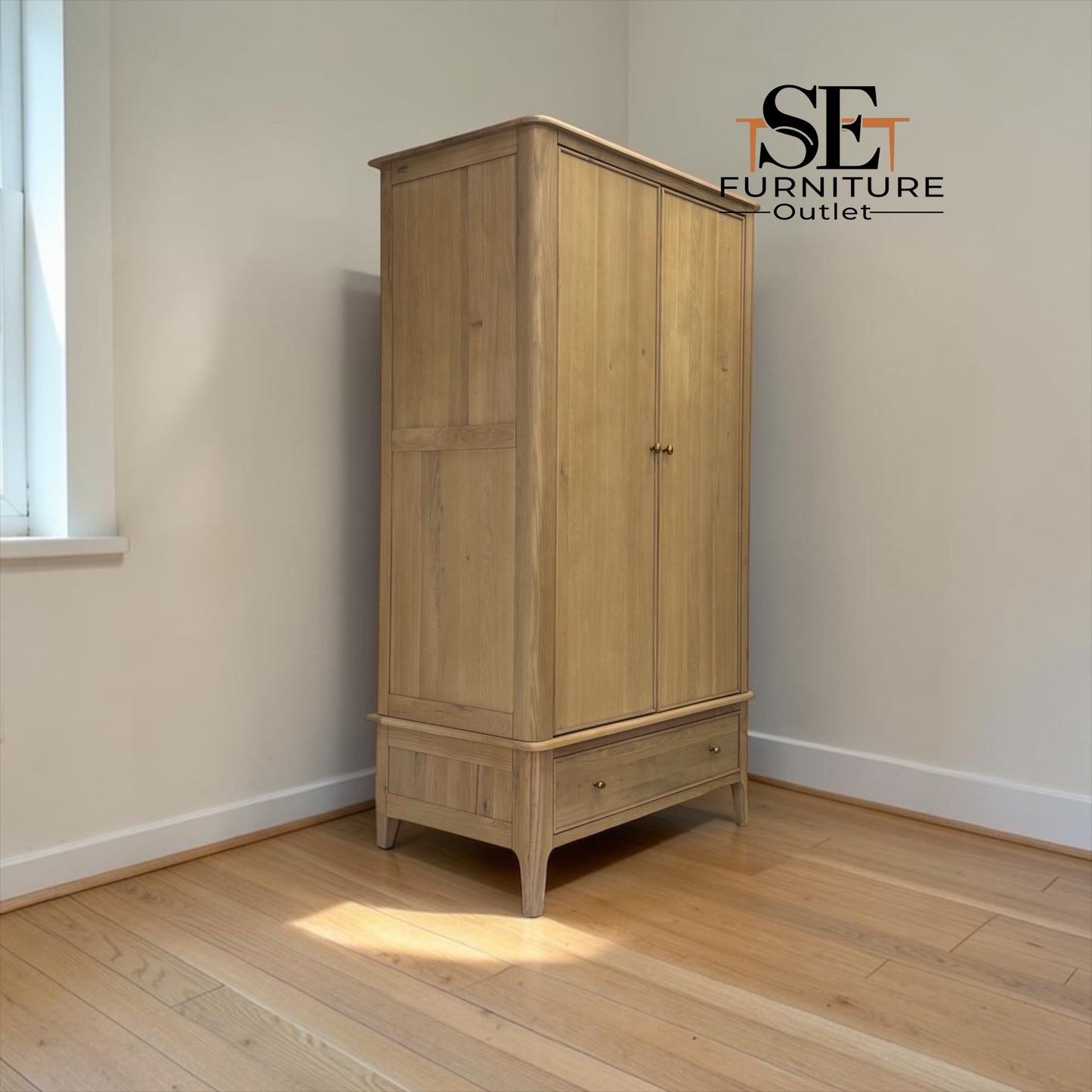 A tall light oak double-door wardrobe with a single drawer, photographed in a minimalist room with beige walls and light wood floors. The SE Furniture Outlet logo is visible in the top right corner.

