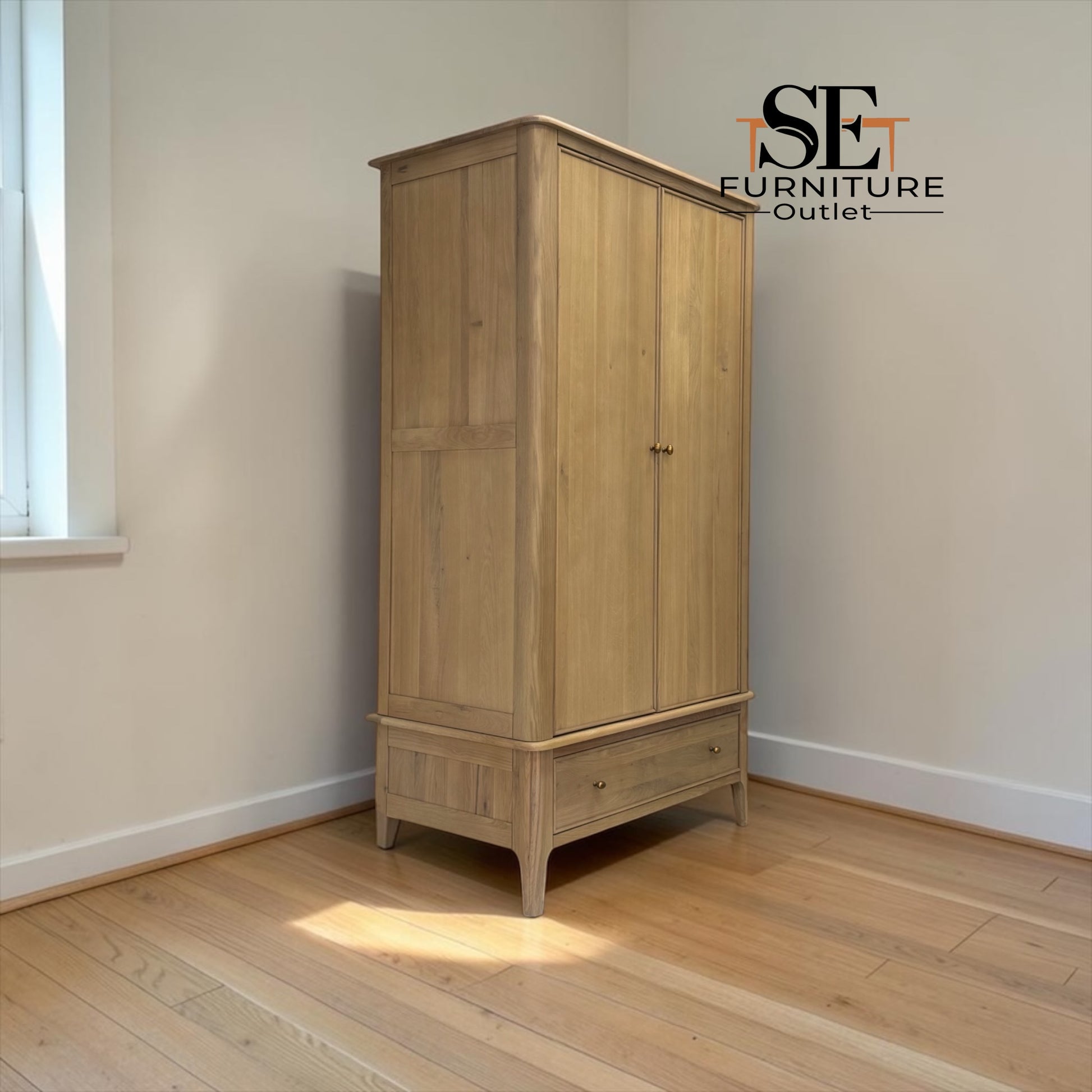 A tall light oak double-door wardrobe with a single drawer, photographed in a minimalist room with beige walls and light wood floors. The SE Furniture Outlet logo is visible in the top right corner.

