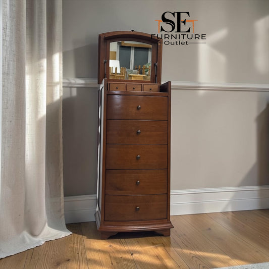 A tall wooden jewelry chest with five drawers and an arched mirror on top, shown in a room with beige walls, a wood floor, and a curtain. The SE Furniture Outlet logo is visible in the upper right.

