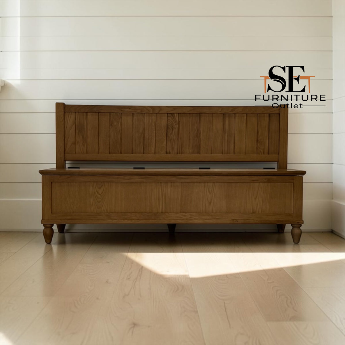 A wooden storage bench with a slatted backrest and turned legs, shown in a bright room with light wood flooring and white shiplap walls. The SE Furniture Outlet logo is visible in the upper right.

