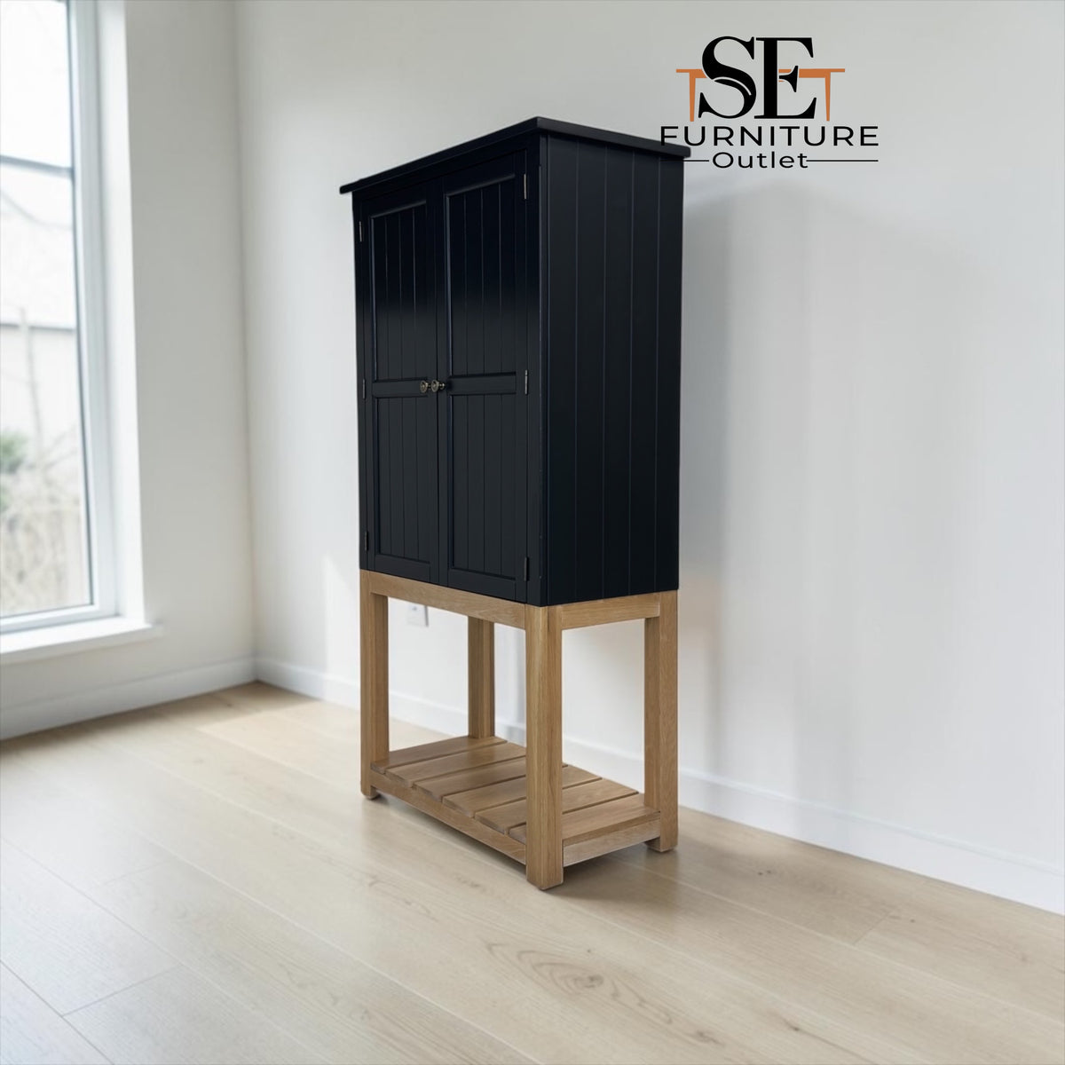 A tall black cabinet with a light natural wood base and open lower shelf, photographed in a bright room with white walls and pale wood flooring. The SET FURNITURE Outlet logo appears in the top right corner.

