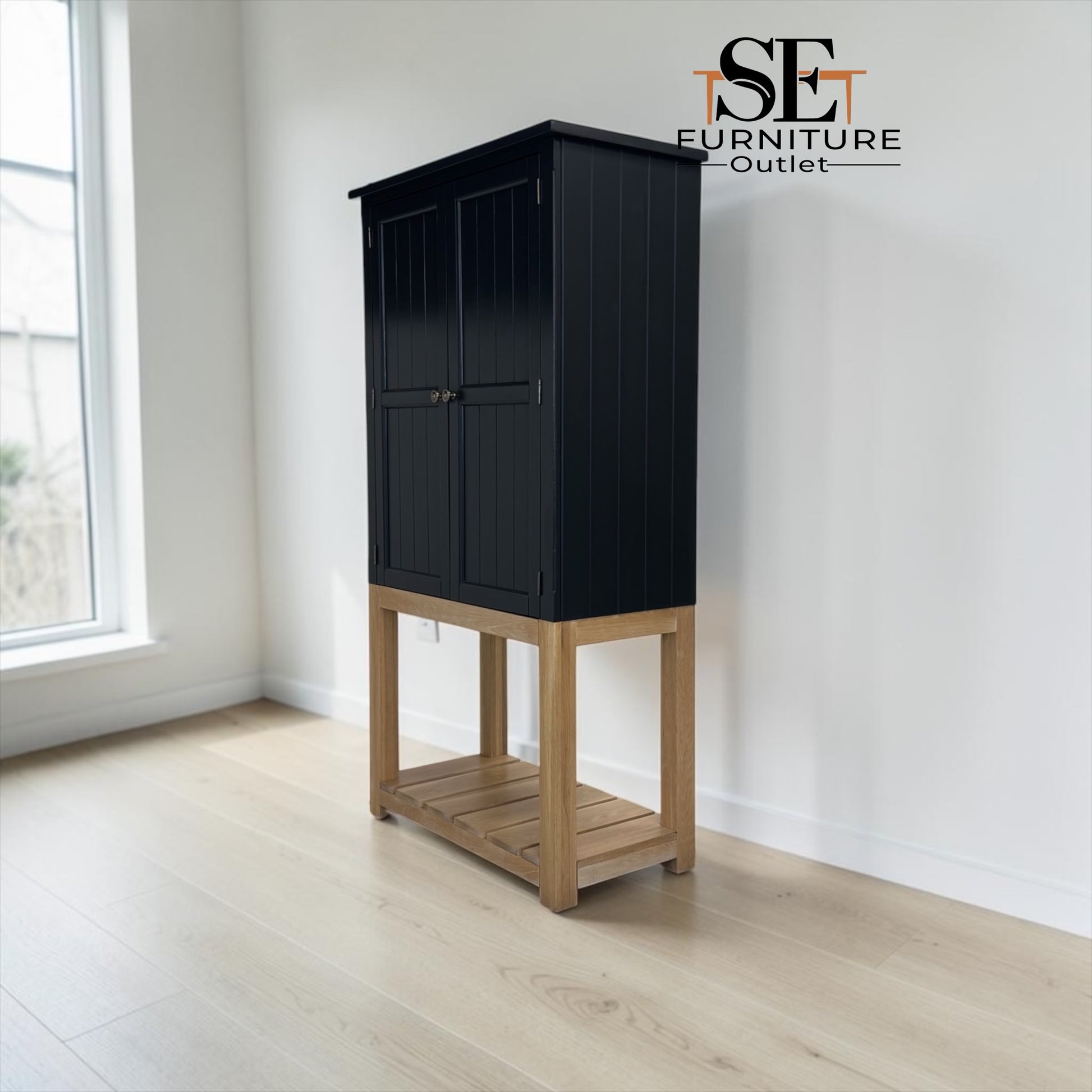 A tall black cabinet with a light natural wood base and open lower shelf, photographed in a bright room with white walls and pale wood flooring. The SET FURNITURE Outlet logo appears in the top right corner.

