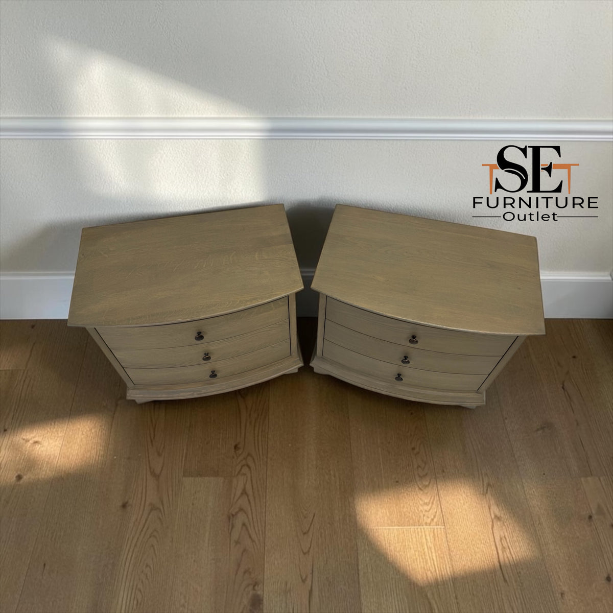 A pair of light beige curved nightstands with three drawers each, placed on a wooden floor against a white wall. The SE Furniture Outlet logo is visible in the top right corner.

