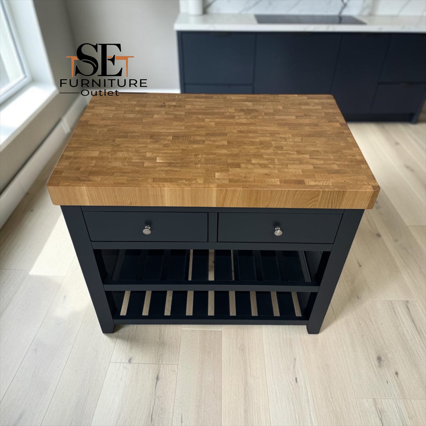 A kitchen island table with a light brown wood butcher block top and a black frame, featuring two drawers with silver knobs and a lower shelf, shown in a bright room with light wood floors.

