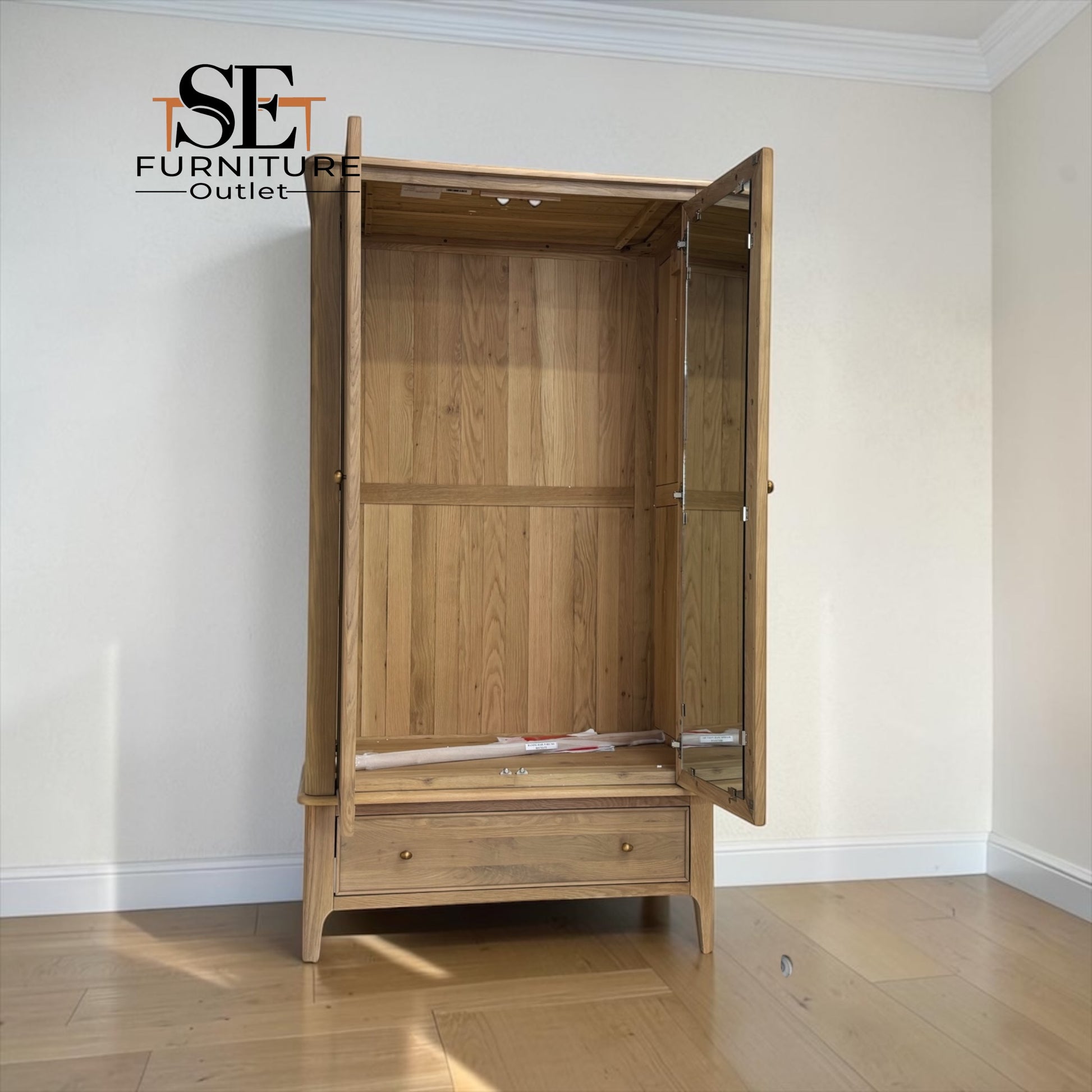 A light oak wood three-drawer wardrobe with mirrored doors, shown open in a room with cream walls and light wood flooring. The 'SE Furniture Outlet' logo is visible in the top left corner.

