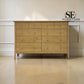 A light oak wood 5-drawer chest of drawers with small round knobs, set against a white paneled wall on a light wood floor. An 'SE FURNITURE Outlet' logo is visible in the top right corner.

