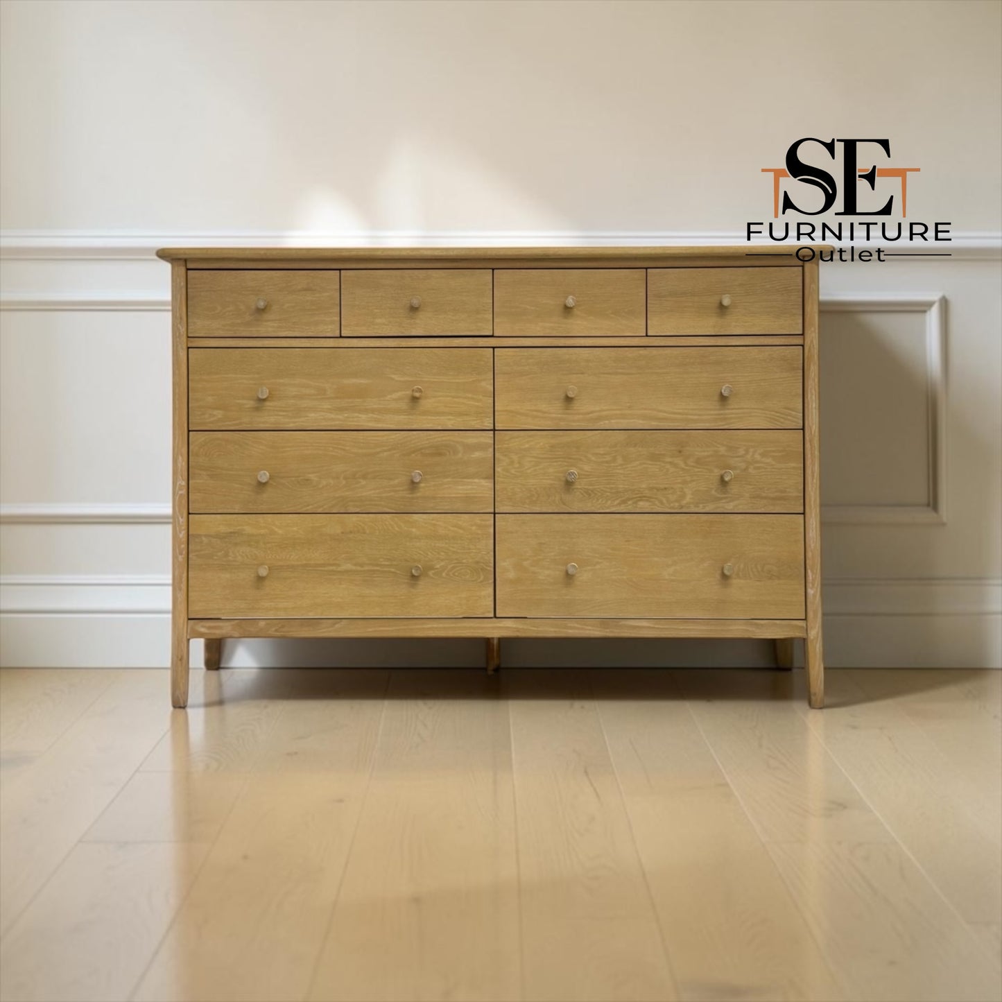 A light oak wood 5-drawer chest of drawers with small round knobs, set against a white paneled wall on a light wood floor. An 'SE FURNITURE Outlet' logo is visible in the top right corner.

