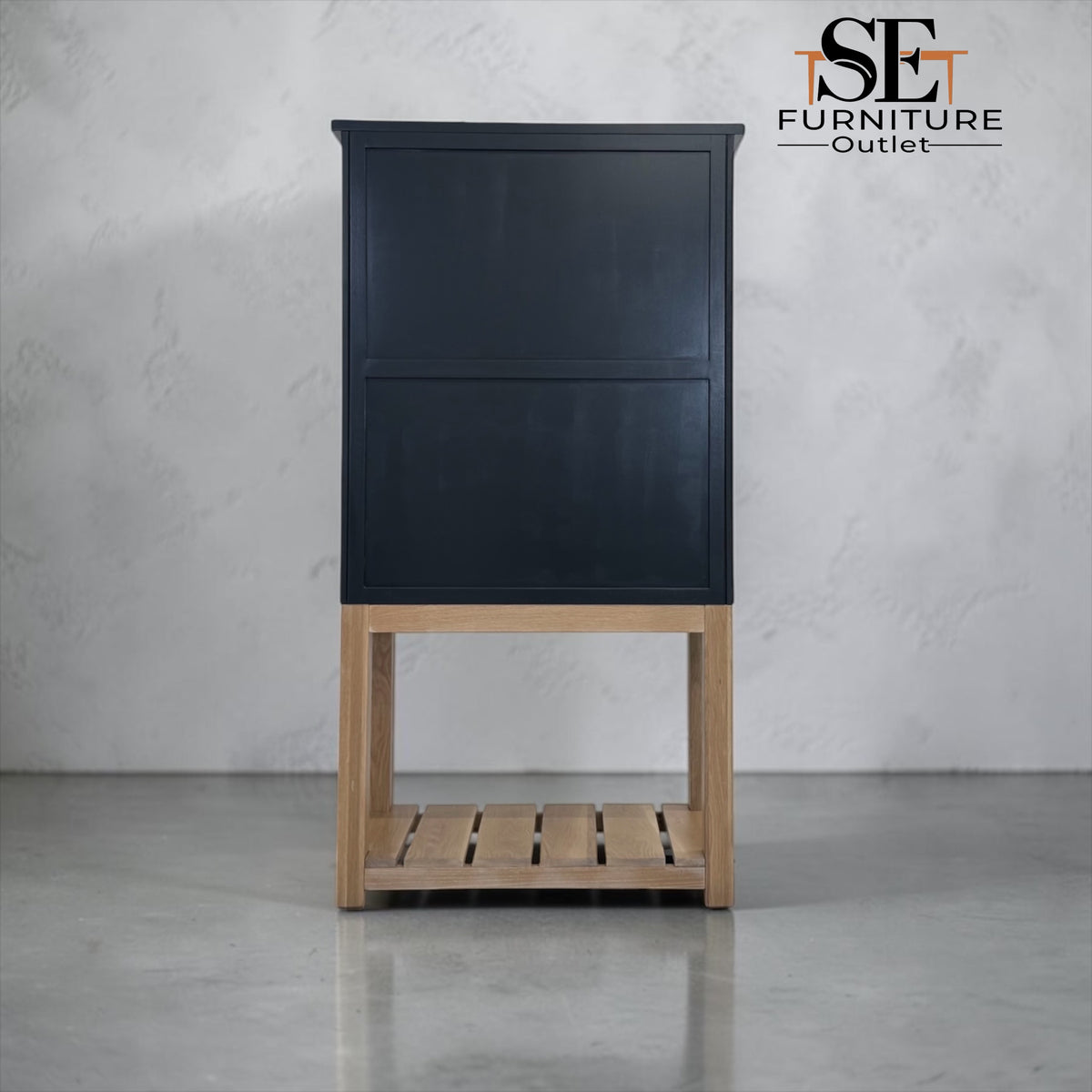 A tall black cabinet with a light natural wood base and open slatted shelf, photographed against a light gray textured wall and floor. The SE Furniture Outlet logo is visible in the top right corner.

