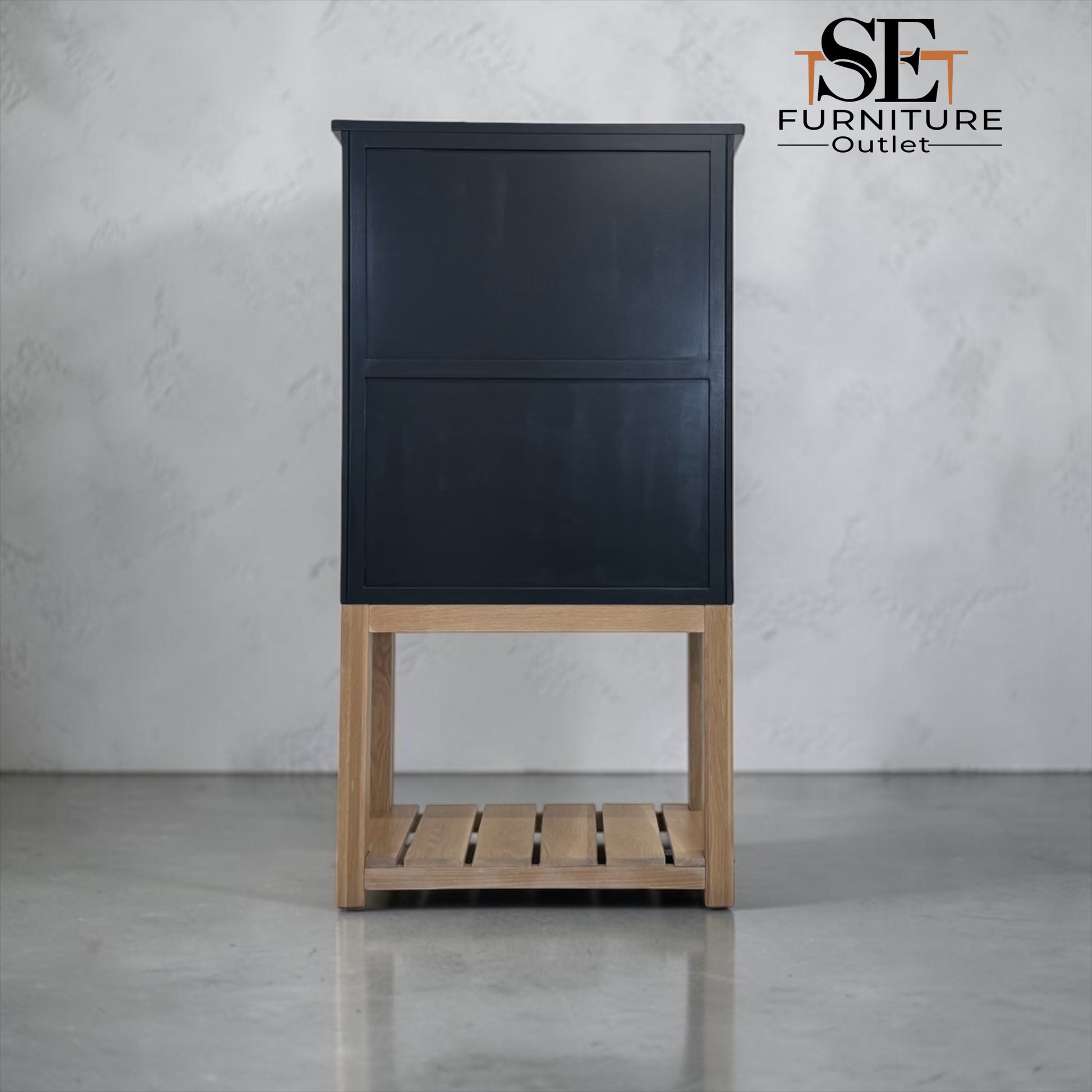 A tall black cabinet with a light natural wood base and open slatted shelf, photographed against a light gray textured wall and floor. The SE Furniture Outlet logo is visible in the top right corner.

