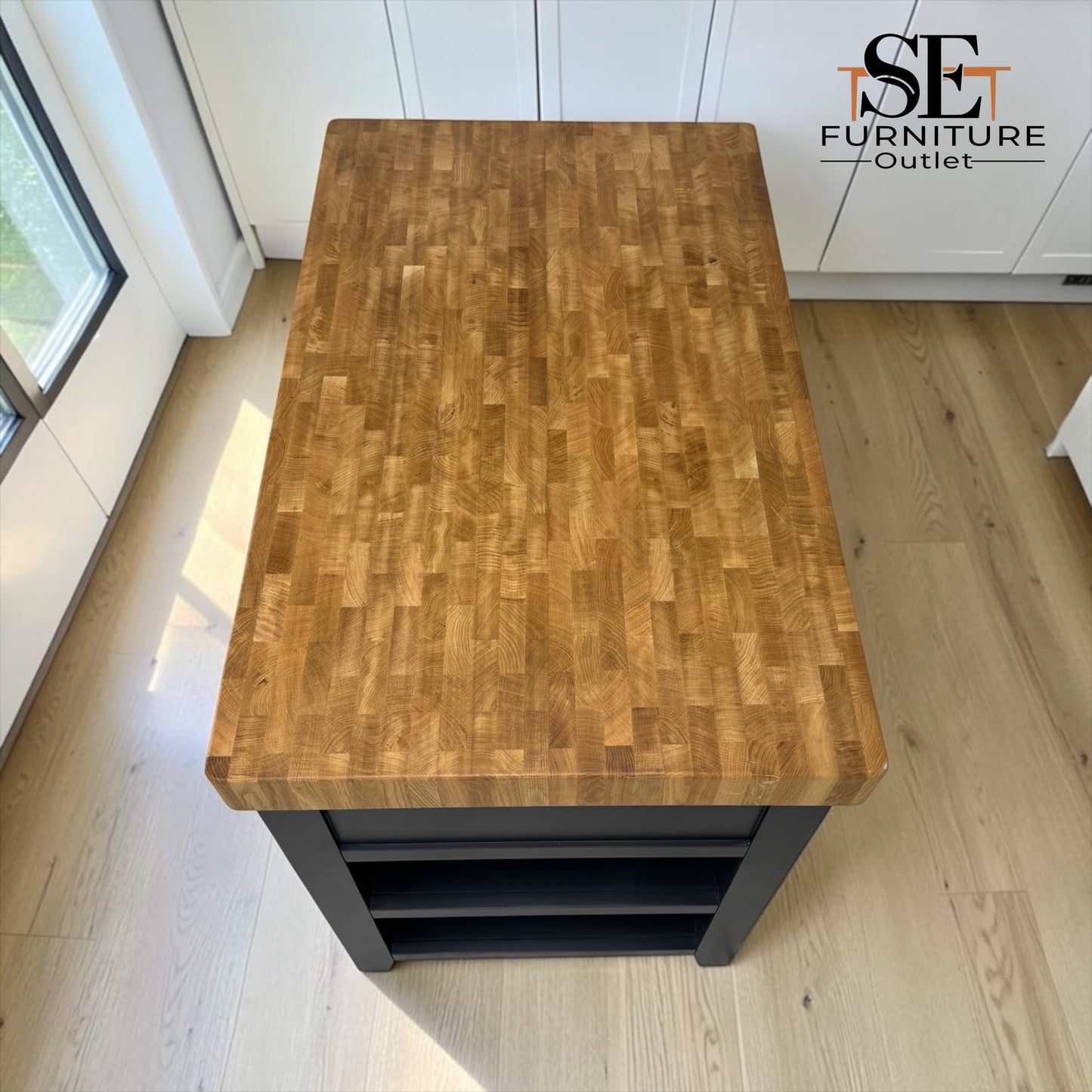 A rectangular butcher block island table with a warm brown wood top and a black metal base, shown in a bright kitchen setting with light wood floors and white cabinets. The 'SE Furniture Outlet' logo is visible in the top right corner.

