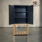 A dark navy blue two-door cabinet with open doors, showing three interior shelves, supported by a light natural wood base with a slatted lower shelf. The photo is taken in a studio-like setting with a neutral beige wall and gray floor, and the SE Furniture Outlet logo is visible in the top right corner.

