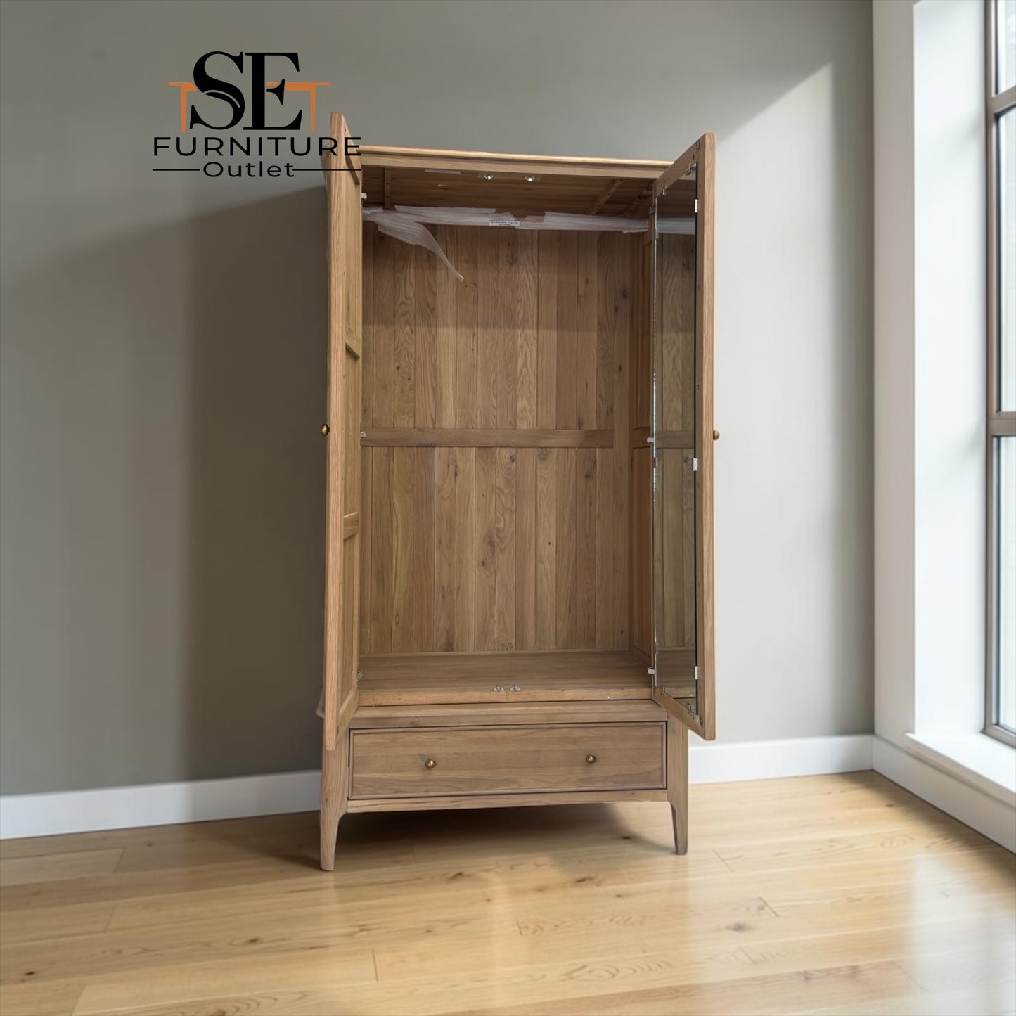 A light oak wood double wardrobe with one door open, showing the interior shelving and glass side panels. The wardrobe is set in a room with light wood floors, a gray wall, and a window on the right.

