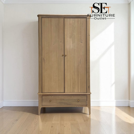 A tall two-door wooden wardrobe or armoire with a single drawer at the bottom, shown in a bright room with light wood flooring and white walls. The cabinet has a natural oak-like finish and small round metal knobs.

