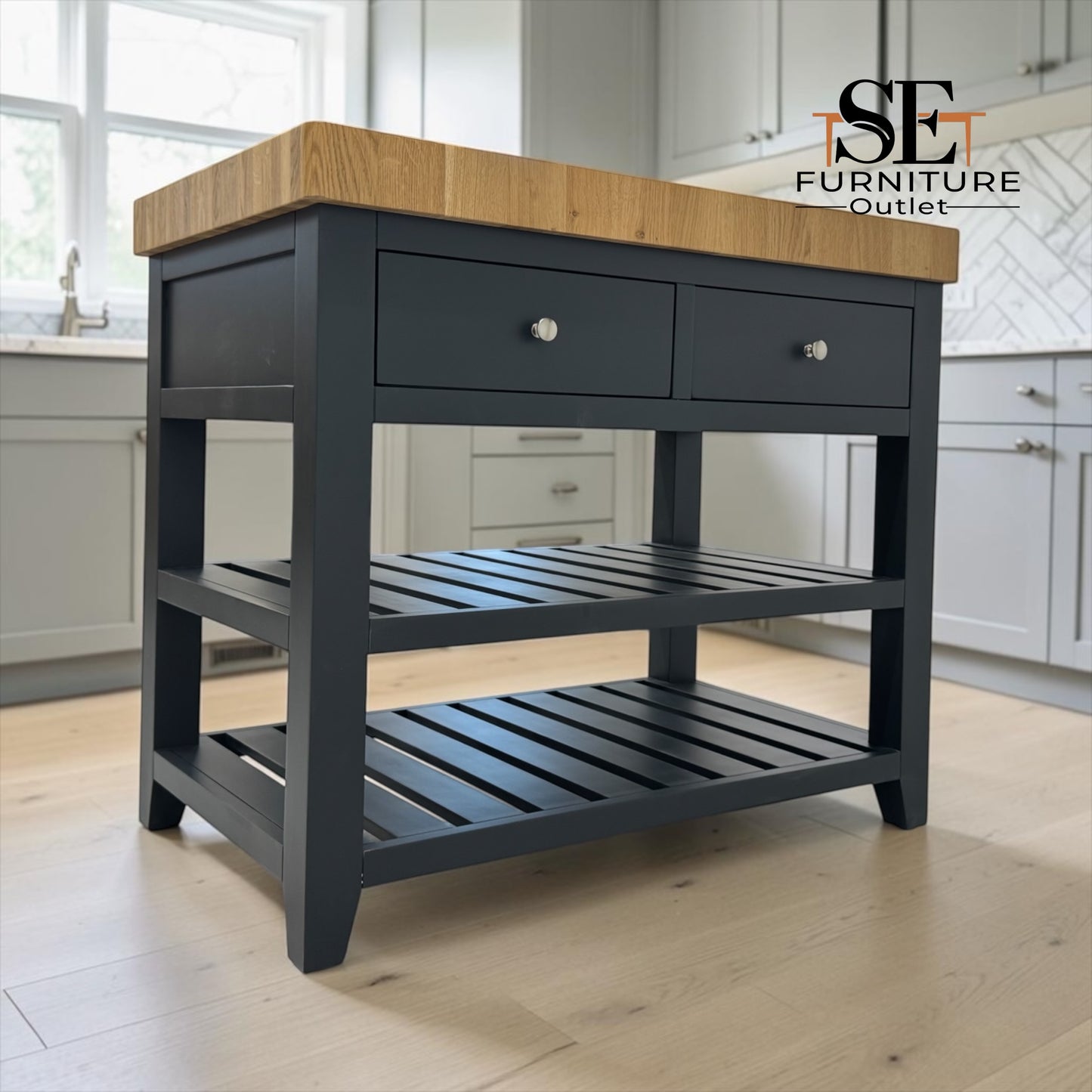 A kitchen island table with a dark charcoal gray frame, two drawers on top, a slatted middle shelf, and a natural oak wood countertop, shown in a bright kitchen setting. An 'SE Furniture Outlet' logo is visible in the top right corner.