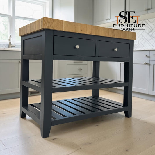 A kitchen island table with a dark charcoal gray frame, two drawers on top, a slatted middle shelf, and a natural oak wood countertop, shown in a bright kitchen setting. An 'SE Furniture Outlet' logo is visible in the top right corner.
