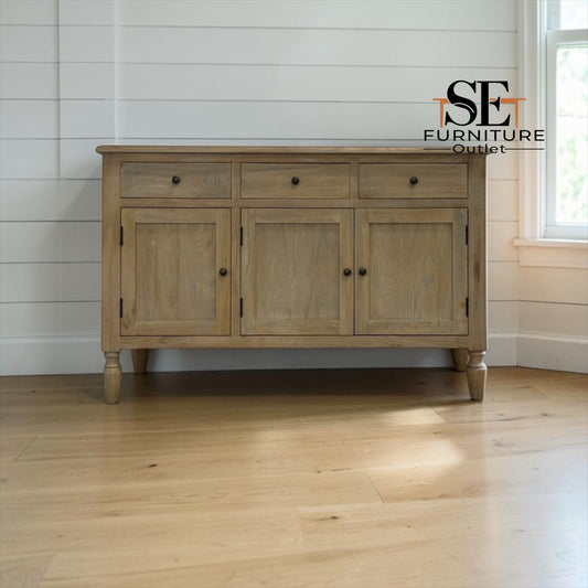 A light oak wood sideboard cabinet with three drawers on top and three paneled doors below, shown in a bright room with white shiplap walls and a wood floor. An 'SE Furniture Outlet' logo is visible in the upper right.

