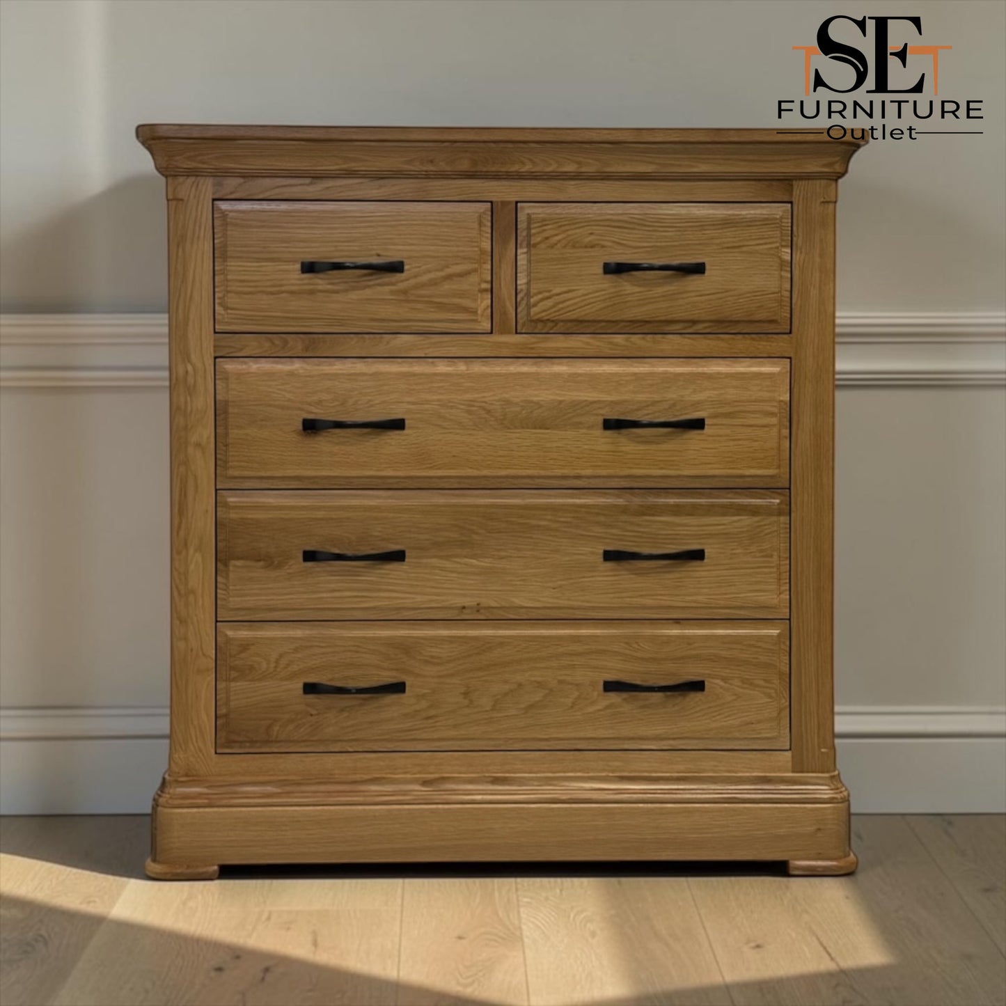 A light oak wood chest of drawers with five drawers and black metal handles, shown in a room setting with a beige wall and light wood floor. An 'SE Furniture Outlet' logo is visible in the top right corner.

