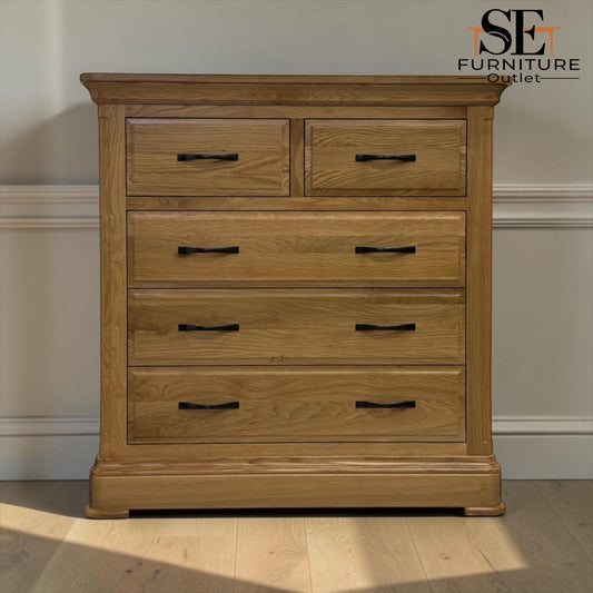 A light oak wood chest of drawers with five drawers and black metal handles, shown in a room setting with a beige wall and light wood floor. An 'SE Furniture Outlet' logo is visible in the top right corner.

