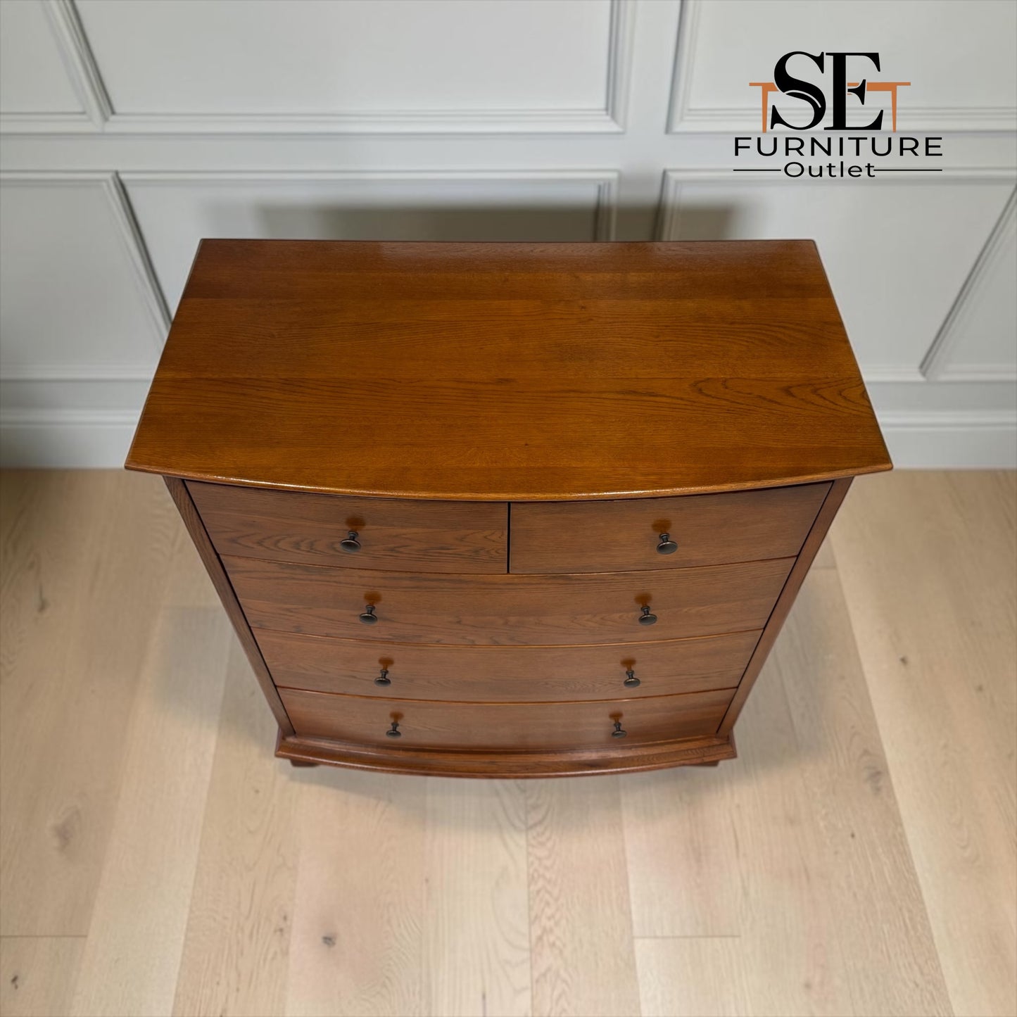 A wooden chest of drawers with a curved front, featuring five drawers with dark metal knobs, set against a white paneled wall and light wood flooring. An 'SE Furniture Outlet' logo is visible in the top right corner.

