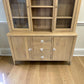 A light natural wood hutch and sideboard cabinet with glass-front upper cabinets, open shelves, two side doors, and three drawers with white decorative pulls, shown on a wood floor against a light wall.


