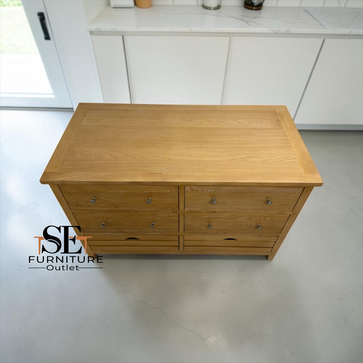 A light oak wood chest of drawers with six drawers and small silver knobs, photographed in a modern kitchen setting. A 'SET FURNITURE Outlet' logo is visible in the bottom-left corner.

