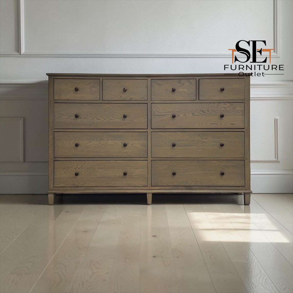 A 12-drawer wooden dresser in a weathered gray-brown finish with small dark knobs, set against a white paneled wall and light wood floor. The SE Furniture Outlet logo is visible in the top right corner.

