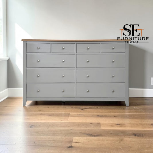 A light gray wooden dresser with ten drawers and small round silver knobs, set against a neutral gray wall with a hardwood floor. The SE Furniture Outlet logo is visible in the top right corner.


