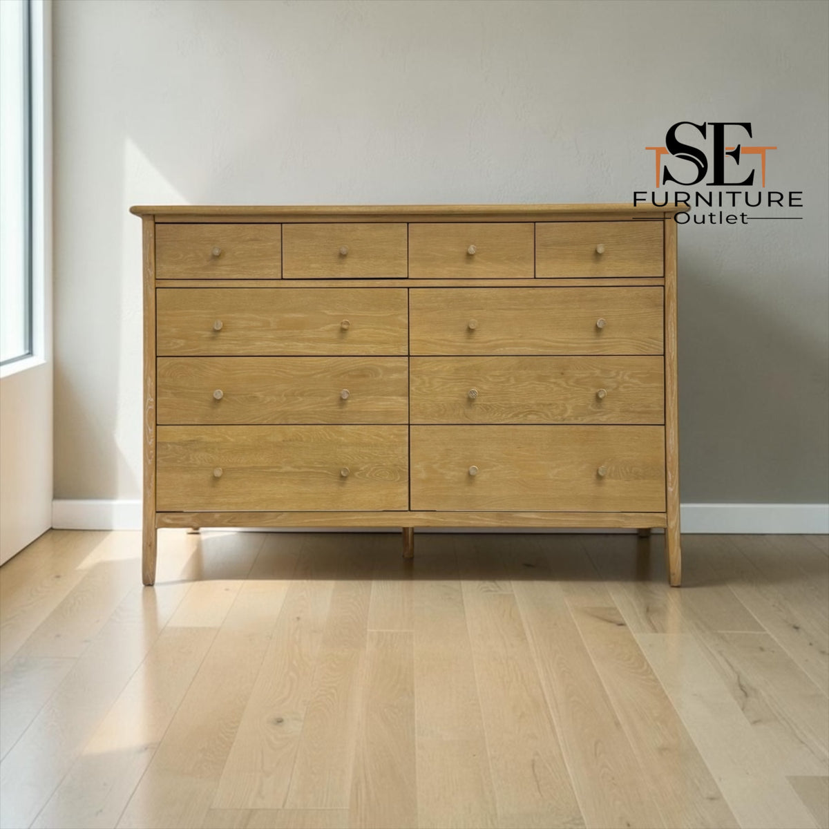 A light oak wood six-drawer dresser with small round knobs, positioned against a neutral gray wall on a light wood floor. The SE Furniture Outlet logo is visible in the top right corner.

