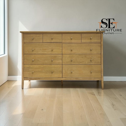 A light oak wood six-drawer dresser with small round knobs, positioned against a neutral gray wall on a light wood floor. The SE Furniture Outlet logo is visible in the top right corner.

